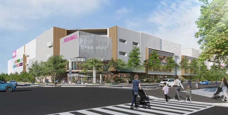 Exterior view of a modern AEON Mall with glass and wood accents, trees, and a busy crosswalk in front.