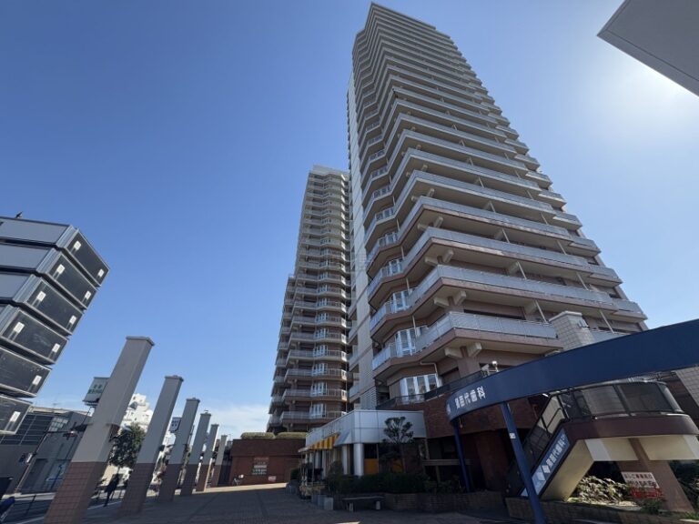 Tall modern apartment building with many balconies rising against a clear blue sky; street-level shops visible at the base.