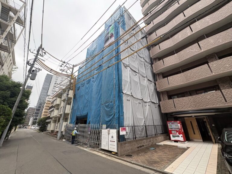 Building under construction wrapped in blue and white tarps along a narrow urban street, with fencing and a worker nearby.
